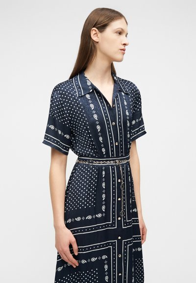 Γυναίκα που φοράει σκούρο μπλε φόρεμα με κουμπιά, κοντά μανίκια, μοτίβα παίζαρι και πουά, και λεπτή αλυσίδα στη μέση, στέκεται μπροστά σε απλό φόντο.