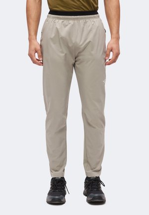 Mand iført lyse beige North Face bukser og sorte vandresko, stående foran en ensfarvet lys grå baggrund.