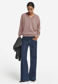 Ljusrosa V-ringad tröja, avslappnad passform, i kombination med mörkblå utsvängda jeans. Modellen håller en grå jacka. Enkel, funktionell design.