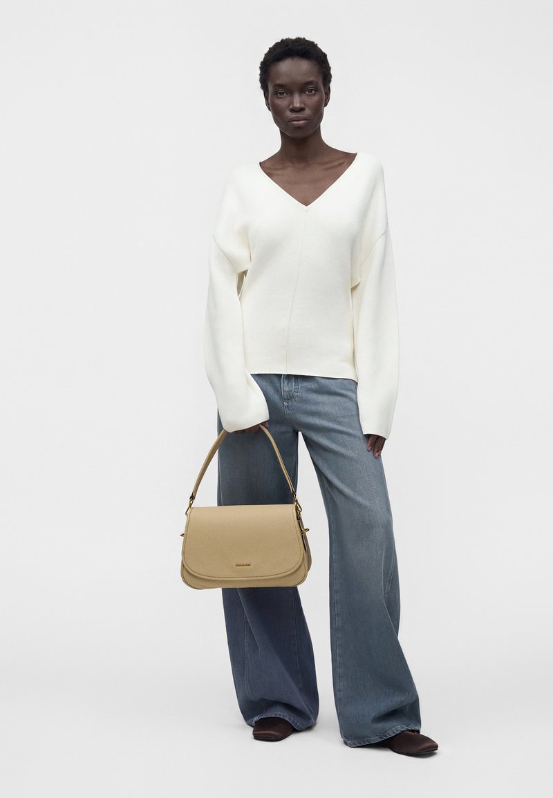 Femme portant un pull blanc col V et un jean à jambes larges, tenant un sac beige avec une petite anse, debout devant un fond uni.
