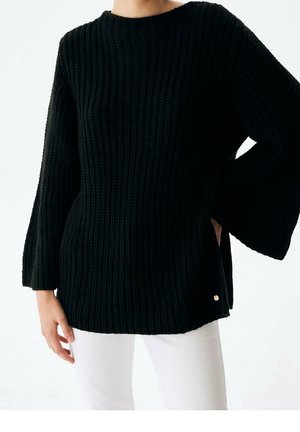 Kvinde iført en sort, ribstrikket sweater med vide ærmer og rund hals, kombineret med hvide bukser foran en ensfarvet baggrund.