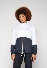 Die Windbreaker-Jacke hat einen weißen oberen Abschnitt und einen dunkelblauen unteren Abschnitt. Sie besteht aus leichtem Material mit elastischen Bündchen und einem gerafften Saum.