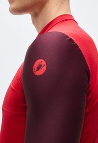 Κοντινό του άνω βραχίονα ενός ατόμου που φοράει κόκκινη και σκούρα μαρόν αθλητική φανέλα με μικρό κόκκινο λογότυπο σκορπιού στο μανίκι.