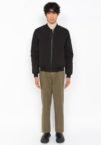 Schwarze Bomberjacke mit einem Reißverschluss vorne, gerippten Bündchen und einer aufgenähten Tasche am Ärmel, kombiniert mit olivgrünen Hosen und schwarzen Schuhen.