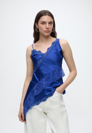 Kvinde iført en blå satin camisole med blondekant og hvide bukser, poserer med den ene hånd i lommen mod en ensfarvet baggrund.