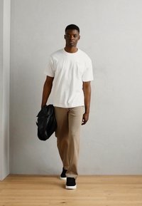 Hvit t-skjorte med rund hals, kombinert med beige bukser og svarte joggesko. Holder en svart veske, går på lys treinnendørsgulv mot en nøytral vegg.