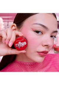 Jeune femme avec maquillage des yeux rose et vernis à ongles tient un pot de blush rouge Maybelline Cloudtopia près de sa joue sur un fond rose.