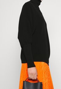 Pull à col roulé noir avec texture côtelée et manches amples, associé à une jupe plissée orange vif et un petit sac à main noir.