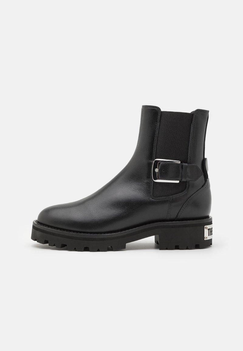 Bottine en cuir noir avec semelle à crampons épaisse, panneau élastique sur le côté et détail boucle argentée sur fond blanc.