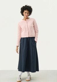 Light pink knitted cardigan with textured fabric, round neckline, and front pockets, paired with a long navy skirt and white sneakers.