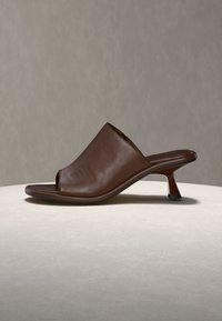 Brown leather open-toe mule with a low curved heel, displayed on a light surface against a plain gray background.