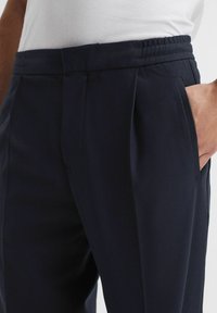 Hombre vistiendo pantalones sastre azul marino oscuro con bolsillos laterales y cintura elástica, mano parcialmente en el bolsillo, combinado con una camisa blanca.