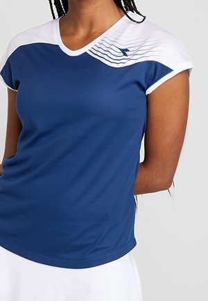 Kobieta ubrana w krótką sportową koszulkę w kolorze niebiesko-białym z małym logo i ukośnym paskiem w okolicy ramienia.