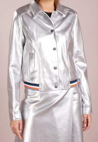 Silver metallic jacka med krage, framknappar och sidofickor. Har ett randigt midjebälte i blått, orange och svart.