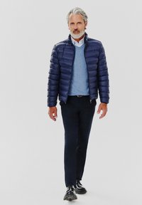 Granatowa puchowa kurtka, lekka, z suwakiem, noszona na jasnoniebieski sweter i koszulę. Ciemne spodnie, czarne buty, ogólnie schludny wygląd.