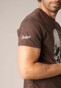 T-shirt en coton marron avec un col rond, des manches courtes et un logo "Deeluxe" blanc sur l'épaule gauche, ainsi qu'un imprimé graphique sur le devant.