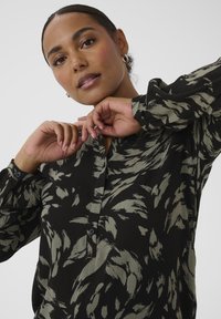 Femme portant un chemisier à manches longues noir et beige à motifs, mains levées près du col, expression faciale neutre, cheveux tirés en arrière.