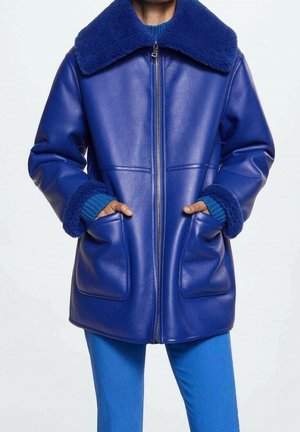 Personne portant une veste en cuir bleue avec col et poignets en laine de mouton, mains dans les poches avant, associée à un pantalon et un pull bleus.