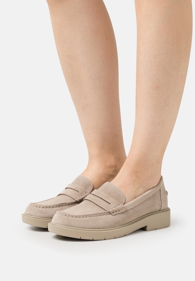 Mocassins en daim beige avec un détail de couture en relief, un bout arrondi et une semelle en caoutchouc beige clair. Texture lisse avec des accents minimaux.