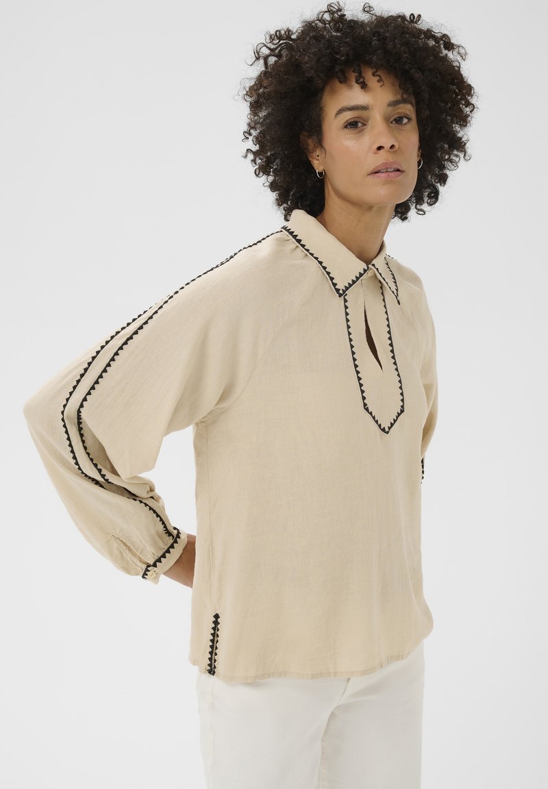 Femme aux cheveux bouclés portant un chemisier beige à manches longues avec un bord en zigzag noir et un pantalon blanc, se tenant devant un fond uni.