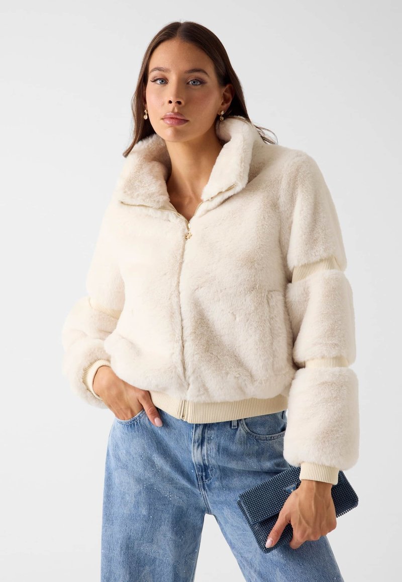 Kremowa kurtka bombowiec z futerkiem z zamkiem błyskawicznym, prążkowanymi mankietami i szerokim kołnierzem; zestawiona z jasnoniebieskimi dżinsami i teksturowaną niebieską kopertówką.