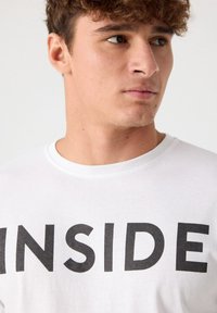 Camiseta de algodón blanca con la palabra "INSIDE" en letras negras y atrevidas en la parte frontal. Diseño clásico de cuello redondo, textura suave y estilo minimalista.