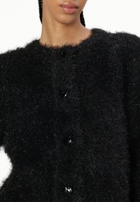 Fekete, bolyhos kardigán texturált, puha felülettel. Kerek nyakkivágás és hat fényes gomb található az elején. Puha és meleg anyag.