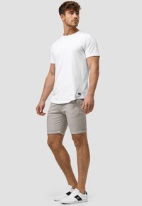 Weißes Baumwoll-T-Shirt und hellgraue Shorts mit umgeschlagenen Säumen. Das Model trägt weiße Sneaker mit schwarzen Akzenten. Neutraler Hintergrund.