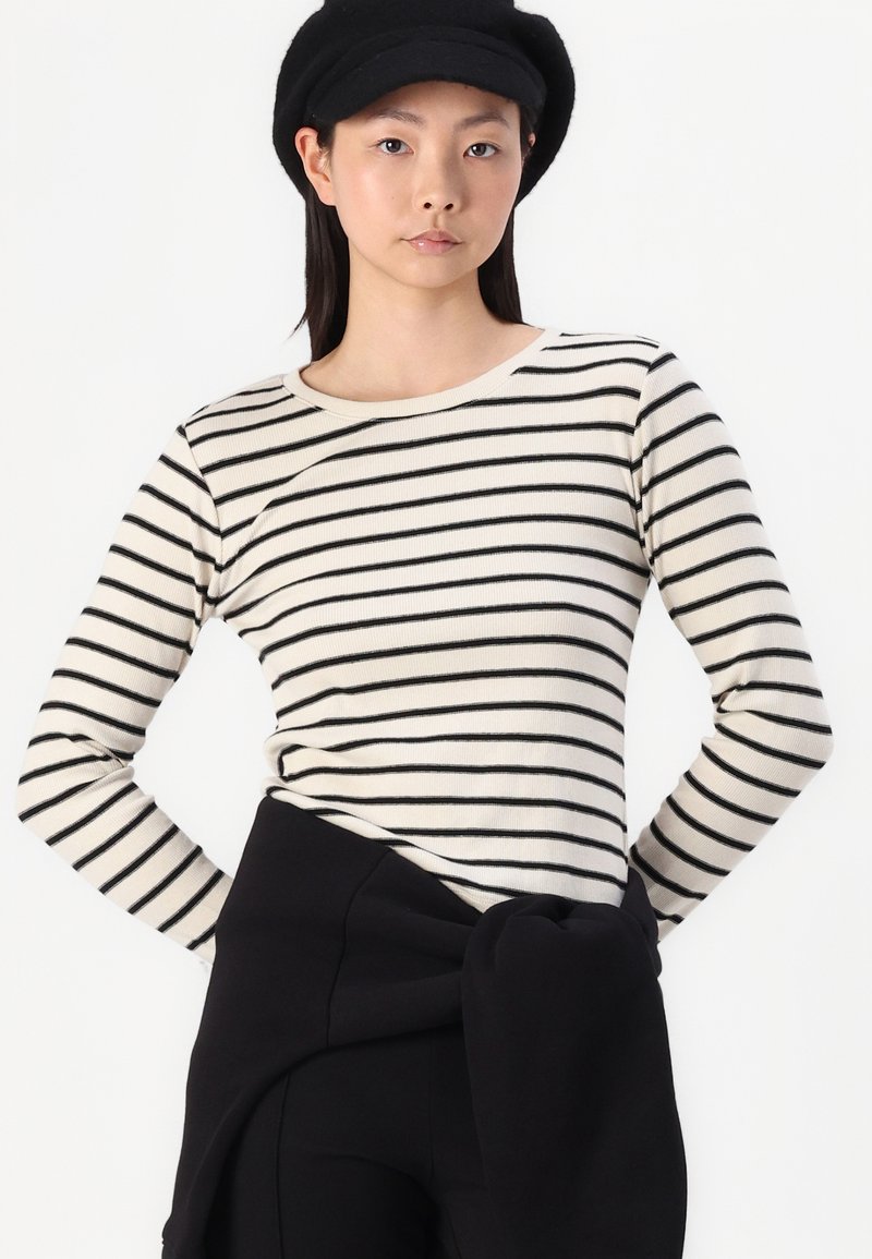 Haut à rayures à manches longues en crème et noir, avec un col rond et une texture côtelée. Associé à un pantalon noir taille haute noué à la taille.