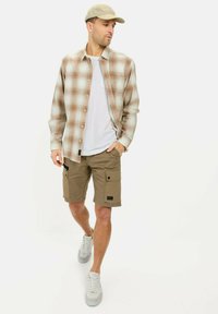 Licht geruite button-up shirt in beige met rode accenten, wit T-shirt, khaki cargo shorts met zakken en grijze sneakers.