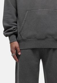 Felpa grigia con tasca frontale e polsini a costine, abbinata a pantaloni coordinati. Tessuto morbido e vestibilità comoda. Anello d'argento alla mano.