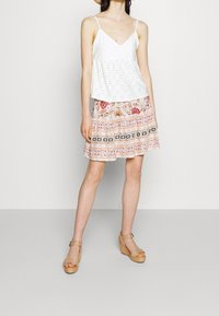 Žena ve bílém bezramenném topu, s vzorovanou růžovo-oranžovou sukní, béžovými platformovými sandály a slaměným kloboukem, stojící na jednobarevném pozadí.