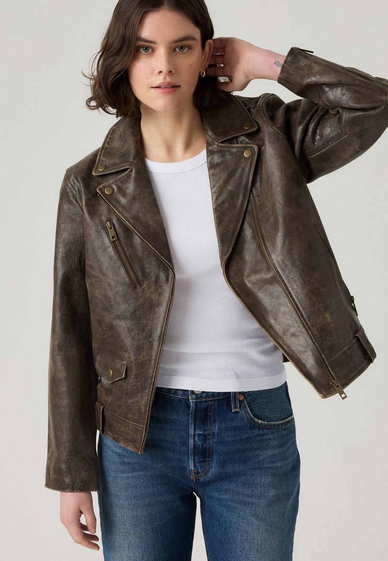 Veste en cuir marron avec des revers angulaires, fermeture éclair à l'avant et poches à boutons. Fini texturé et design courts. Portée par-dessus un haut blanc.