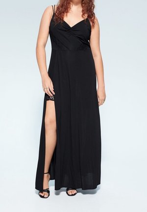 Femme portant une robe maxi noire sans manches avec une fente haute sur la cuisse et des sandales noires à lanières, debout devant un fond clair.