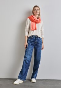 Jeans blu a gamba larga, maglione beige e vivace sciarpa corallo; sneakers bianche. Tessuto denim liscio; morbido tessuto a maglia.