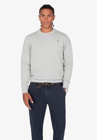 Sudadera de punto gris con cuello redondo, que presenta un pequeño logo azul en el pecho. Combinada con pantalones azul marino oscuros y un cinturón trenzado.
