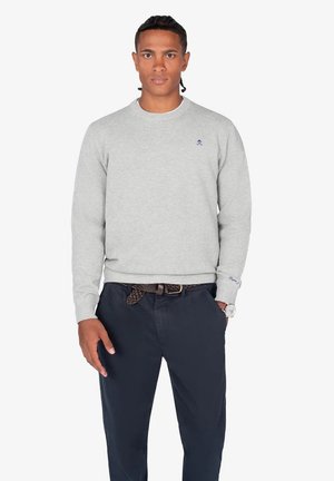 Sudadera de punto gris con cuello redondo, que presenta un pequeño logo azul en el pecho. Combinada con pantalones azul marino oscuros y un cinturón trenzado.