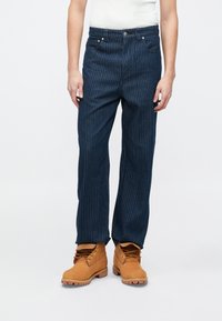 Σκούρα τζιν παντελόνια με κάθετες ανοιχτόχρωμες ρίγες, με κλείσιμο με κουμπί, θηλιές για ζώνη και ίσια γραμμή στο πόδι, συνδυασμένα με καφέ εργασιακά μποτάκια.