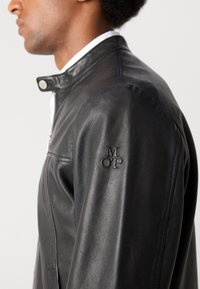Schwarze Lederjacke mit Reißverschluss und Druckknopfkragen. Mit eingeprägtem "M OP"-Logo auf der linken Schulter. Glatte Textur und taillierte Passform.