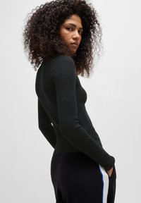 Zwarte geribbelde blouse met lange mouwen, een aansluitende pasvorm en hoge halslijn, gecombineerd met zwarte broeken met witte en blauwe zijstrepen.