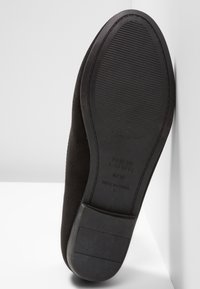 Semelle plate noire de chaussure avec grip texturé et talon bloc, reposant verticalement contre une surface d’angle blanche.