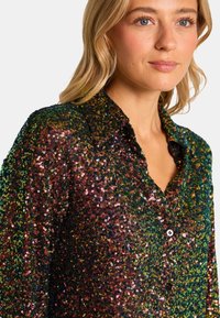 Camisa brillante hecha de lentejuelas multicolores en verde, marrón y dorado, con cuello y cierre de botones. Textura suave visible.