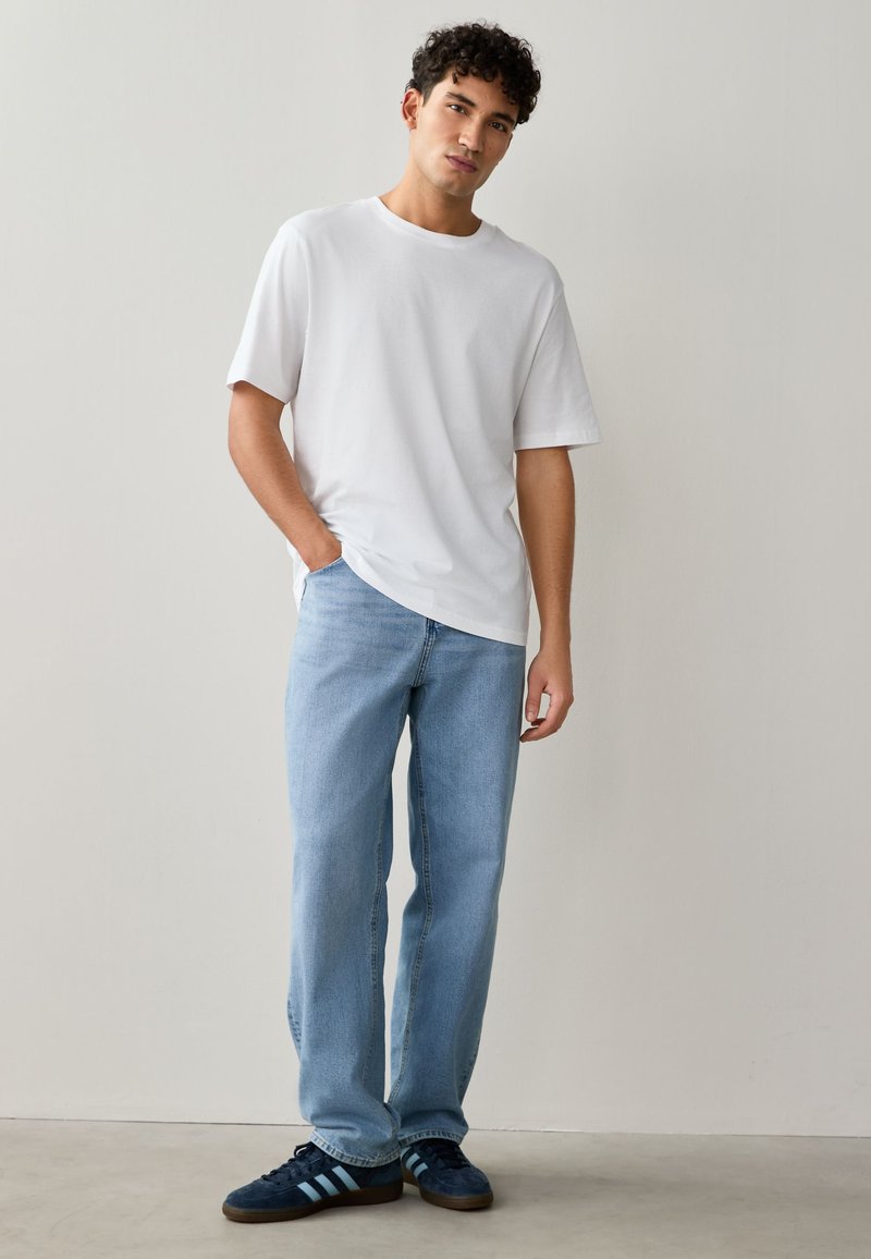 Jeune homme portant un t-shirt blanc uni, un jean bleu clair et des baskets bleu foncé, debout contre un mur clair uni.