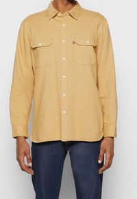 Chemise à manches longues de couleur moutarde en tissu doux, dotée d'une patte de boutonnage à l'avant, d'un col et de deux poches poitrine avec rabats.