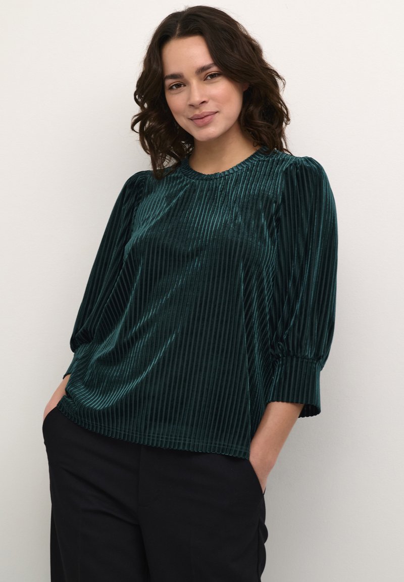 Top de terciopelo acanalado en color azul verdoso con un escote redondo, mangas tres cuartos y detalles fruncidos en los hombros. Combinado con pantalones negros.