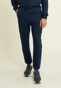 Uomo che indossa pantaloni jogger blu navy con coulisse in vita e sneakers grigio scuro, in piedi con una mano in tasca.