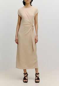 Beiges, knielanges Kleid mit einem verdrehten Knoten-Detail an der Taille, kurzen Ärmeln und einer glatten Textur. Getragen mit schwarzen, schmalen, hohen Sandalen.
