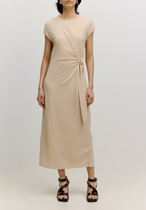 Beige, knielange jurk met een gedraaide knoopdetail bij de taille, mouwen tot de schouder en een gladde textuur. Gekozen met zwarte, gespsandalen met hakken.
