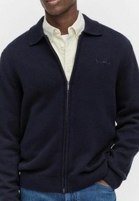 Man som bär en marinblå zip-up tröja över en krämfärgad skjorta med knappar, med ena handen delvis i fickan, visas från bröstet till mitt lår.