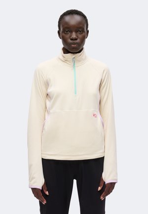 Femme portant un pull beige clair à manches longues avec une fermeture éclair au quart, et un pantalon noir, debout devant un fond uni.